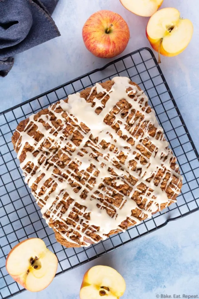 An apple coffee cake with a crumb topping and a salted caramel glaze on a cooling rack with apples.