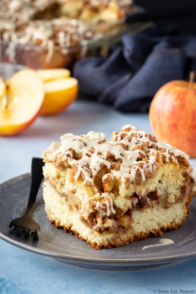 A piece of apple coffee cake on a plate with apples and the pan of coffee cake.
