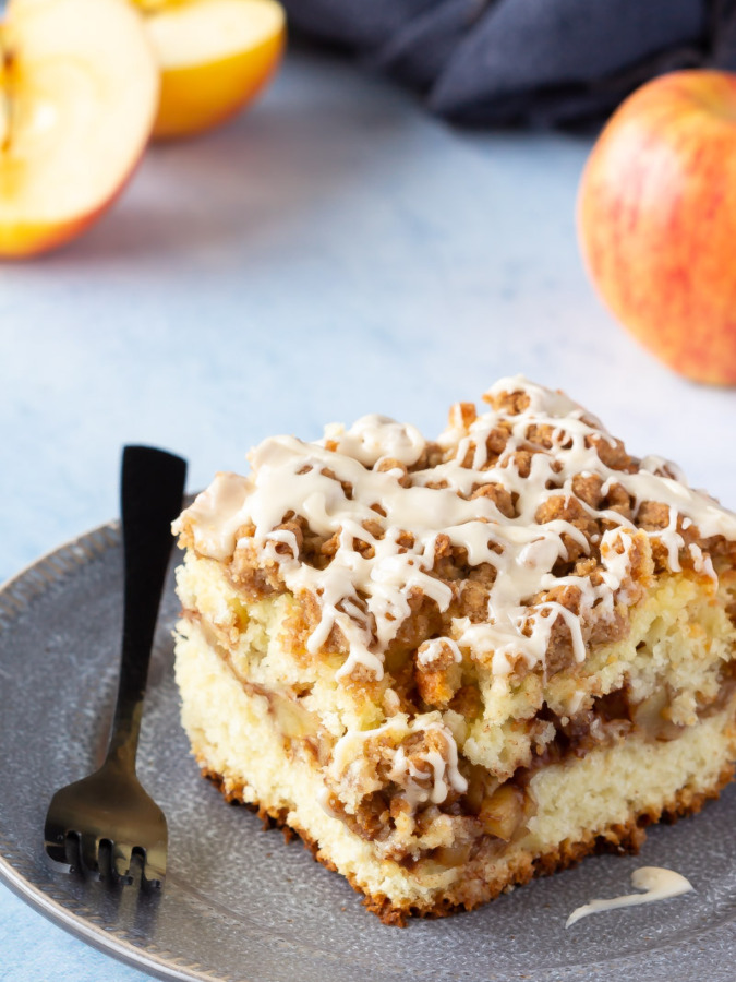 Piece of apple cinnamon filled coffee cake with a crumb topping and a salted caramel glaze on a place with apples.