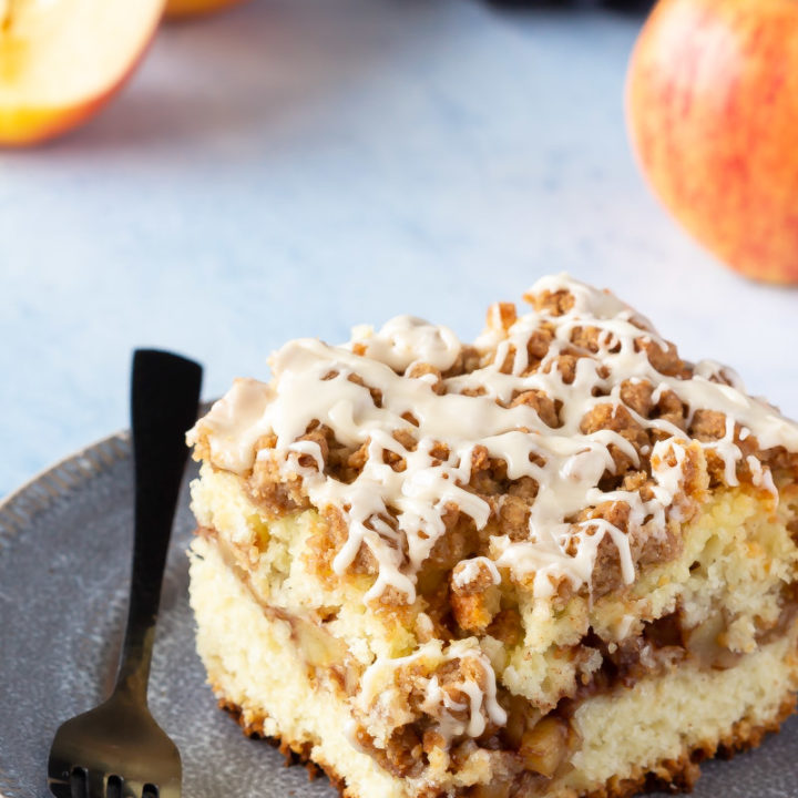 Piece of apple cinnamon filled coffee cake with a crumb topping and a salted caramel glaze on a place with apples.